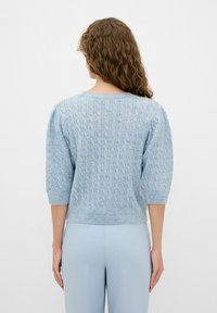 Maglione lavorato a maglia di colore azzurro chiaro con un motivo a trecce testurizzato, maniche a sbuffo corte e orlo a costine, indossato sopra pantaloni abbinati di colore azzurro chiaro.
