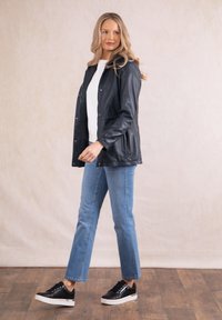 Black leather jacket with snap buttons, white top, light blue jeans, and black sneakers with white soles, set against a neutral background.