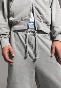 Persona che indossa pantaloni della tuta grigio chiaro con coulisse e felpa con cappuccio abbinata con zip, fascia della biancheria intima bianca e blu visibile sotto la felpa parzialmente aperta.