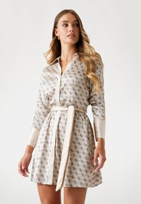 Femeie îmbrăcată cu o rochie bej cu model, până la genunchi, cu o curea, mâneci lungi și guler, stând în fața unui fundal simplu, deschis la culoare.