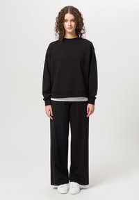 Černá mikina s dlouhými rukávy a kulatým výstřihem, doplněná volnými černými kalhotami. Bílé tenisky dotvářejí vzhled. Měkká textilní struktura.