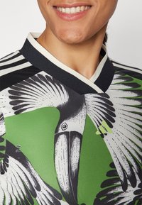 Kortärmad skjorta med en färgglad fågelfeldesign i svart och grönt, med svart krage och randiga detaljer, tillverkad av ett mjukt tyg.