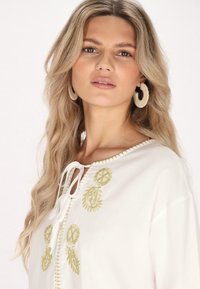 Weiße Bluse mit grünen bestickten Akzenten und einer Bindeschließe am Halsauschnitt; verfügt über einen runden Kragen und eine weiche Stoffstruktur.