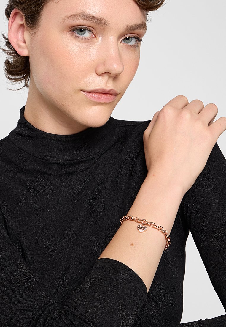 Eine roségoldene Gliederarmband mit Herzanhänger, bestehend aus ineinandergreifenden Gliedern und einer polierten Oberfläche, getragen an einem Handgelenk über einem schwarzen Rollkragenpullover.