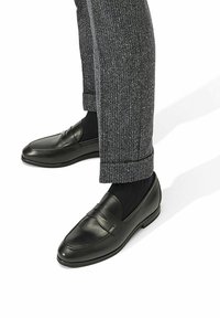 Zwarte leren instappers met een gladde afwerking en decoratieve stiksels, gedragen met grijze gestructureerde broek en zwarte sokken.