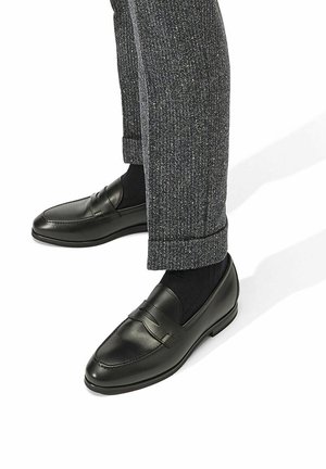 Mocasines de cuero negro con un acabado liso y costuras decorativas, usados con pantalones grises de textura y calcetines negros.