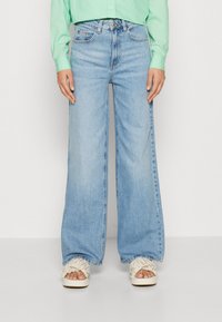 Jeans de pierna ancha de color azul claro con cintura alta, cinco bolsillos y un ligero desgaste. Combinados con zapatos de plataforma color crema con detalles de volantes.