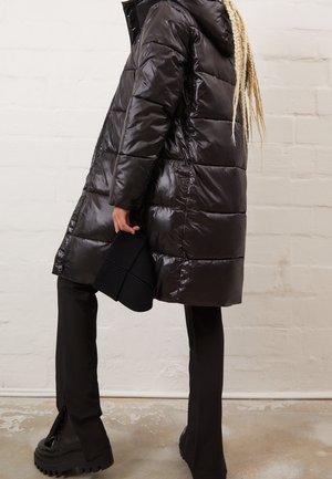 Personne avec de longues tresses blondes portant un manteau matelassé noir, un pantalon noir, des bottines à plateforme, tenant un bonnet en tricot noir contre un mur de briques blanches.