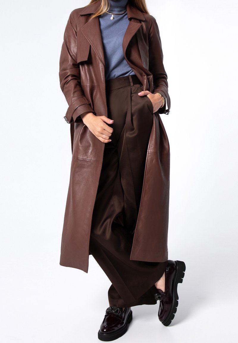 Femme portant un long manteau en cuir marron, un col roulé bleu, un pantalon large marron et des mocassins noirs, debout avec une main dans la poche.