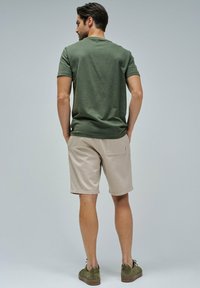 T-shirt de algodón de color verde oscuro, pantalones cortos caqui y zapatillas de color verde oliva. El modelo está de pie con las manos en los bolsillos, mostrando un estilo y ajuste casual.