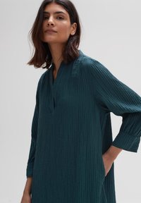 Türkisfarbenes Langarmkleid mit V-Ausschnitt, vertikaler Rippstruktur und seitlichen Taschen. Weicher, leichter Stoff für hohen Tragekomfort.