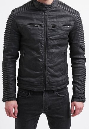 Homme portant une veste en cuir noire cintrée avec des détails côtelés aux épaules et aux manches, associée à un pantalon noir.