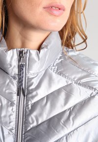 Silverfärgad isolerad jacka med hög krage, dragkedjestängning och chevronmönster med slät, glansig yta.