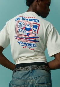 Hvid bomulds t-shirt med røde og blå sushi-grafikker på ryggen. Kombineret med blå denim jeans og et nitter-bælte i sort.