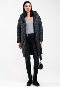 Abrigo largo acolchado negro, camiseta sin mangas gris, jeans ajustados negros, botas de tobillo negras; los accesorios incluyen un bolso pequeño y un collar de cadena de plata.