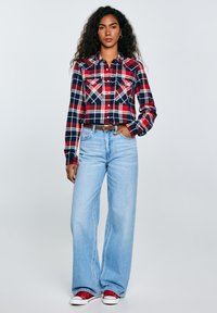 Woman with long curly hair wearing a red and navy plaid shirt, light blue wide-leg jeans, brown belt, and red sneakers, standing with one hand in pocket.