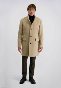 Beige Wollmantel mit hohem Kragen, Knopfverschluss und zwei vorderen Taschen. Getragen über einem schwarzen Rollkragenpullover und kombiniert mit dunkel gemusterten Hosen.