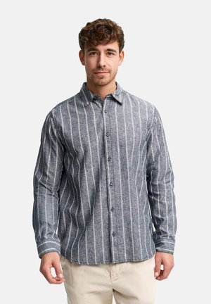 Młody mężczyzna o kręconych włosach, ubrany w szarą koszulę w pionowe paski na guziki oraz jasnobeżowe spodnie, stojący i zwrócony przodem.