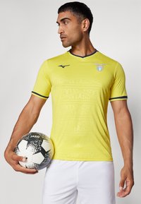 Dzeltena īsām piedurknēm futbolista krekle ar smalkiem teksta rakstiem un zilu apkakli. Melnbaltais futbols tiek turēts kreisajā rokā.