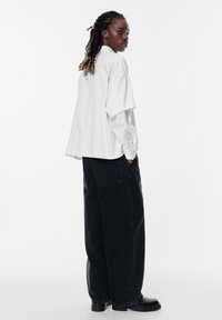 Witte gestreepte oversized shirt met een hoge hals en korte mouwen, gecombineerd met losse zwarte jeans en zwarte schoenen.