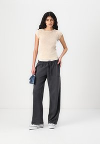 Light beige fitted top with short sleeves, paired with loose dark gray sweatpants. The model holds a small blue bag and wears white sneakers.