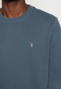 Ciemnoniebieska bluza z bawełny o miękkiej fakturze, z prążkowanym dekoltem typu crewneck oraz małą białą haftowaną czaszką kozy na lewej piersi.