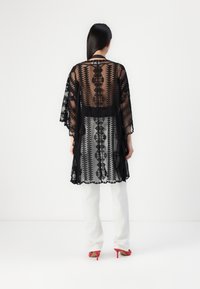 Svart genomskinlig kimono med intrikata geometriska mönster och scallopade kanter, matchad med vita byxor och röda högklackade skor.