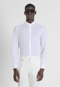 Camisa blanca de manga larga hecha de tela ligera, con un cuello clásico y cierre frontal de botones. Combinada con pantalones de color claro.