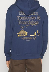 Retro con scritta gialla "Harajuku Teahouse & Boardshop, Aperto su richiesta," e illustrazione di una casa tradizionale e alberi.