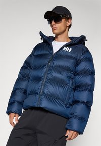 Doudoune bleu marine avec fermeture éclair à l'avant, capuche et sections rembourrées, arborant un logo blanc sur la poitrine. Portée avec un pantalon noir et un chapeau.