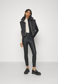 Fekete puffervest magas gallérral, egy krém színű bordás pulóver felett, magasan derekú fekete skinny farmernadrággal és vastag fekete loaferekkel párosítva.