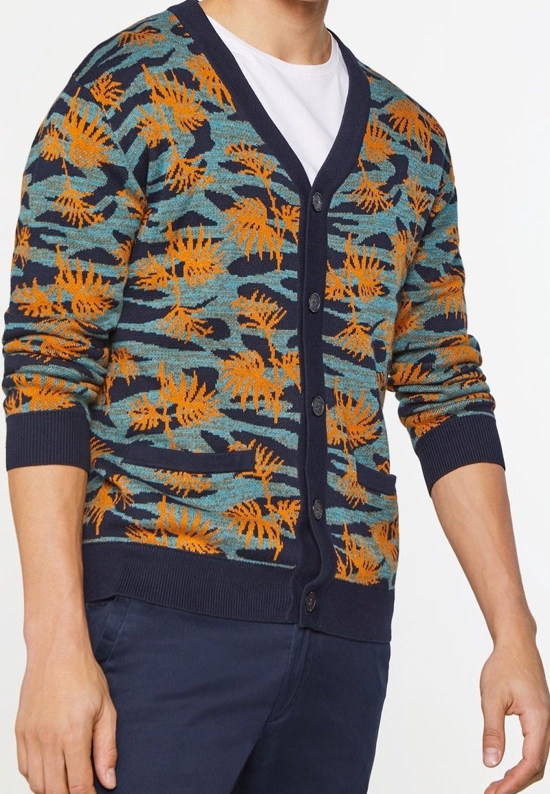 Knappup cardigan med en marinblå bas, med ett mönster av orange och turkosa ormbunkar. Stickad textur med ribbade ärmslut och fickor.