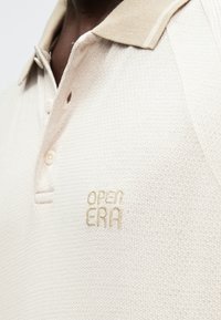 Smėlio spalvos polo marškinėliai iš tekstūruotos medžiagos, su šviesiai rudos spalvos apykakle, trimis mygtukais ir "OPEN ERA" logotipu tokiu pačiu siuvinėjimu.