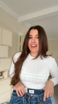 Top de manga larga blanco y acanalado combinado con jeans de mezclilla azules, mostrando la cinturilla y la etiqueta. El fondo presenta muebles de color claro.