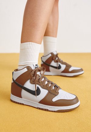 Henkilö, joka käyttää ruskeavalkoisia Nike'n korkeavartisia lenkkitossuja mustalla swoosh-logolla ja valkoisia ribbineuleisia keskipitkiä sukkia keltaisella lattialla.