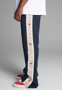 Navy sweatpants with cream side stripes featuring "SIK SILK" logo in red and gold. Zippered cuffs; cotton material. Worn with red sneakers.
