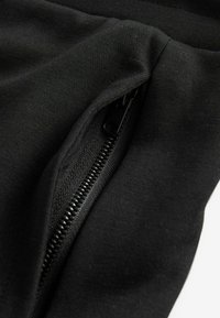 Primer plano de una tela negra con un bolsillo con cremallera. La cremallera es negra con costura texturizada alrededor de la abertura. Textura suave en general.