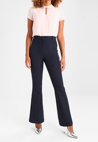 Mujer vistiendo una blusa de manga corta rosa clara, pantalones acampanados de talle alto en color azul marino y tacones altos puntiagudos plateados, de pie contra un fondo blanco.