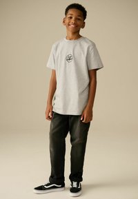 T-shirt gris à manches courtes avec un logo noir circulaire, associé à un jean foncé et des baskets noires avec des accents blancs. Texture de tissu lisse.