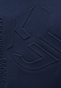 Námornícka modrá textília s embosovaným logom a textom. Hladký povrch s jemným leskom. Jednoduchý, minimalistický dizajn zdôrazňuje značku.