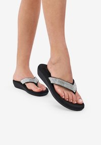 Infradito neri con una suola testurizzata, dotati di un cinturino lucido decorato con strass a forma di V che attraversa il piede.