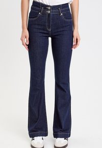 Ciemnoniebieskie dżinsy typu dzwony z zapięciem na dwa guziki, pięcioma kieszeniami, kontrastowym przeszyciem i delikatnym przetarciem na końcu nogawki. Noszone z białymi trampkami.