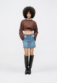 Rjava obrezana jopa z vezenim detajlom, kombinirana s svetlo modro jeans mini krilom s sprednjimi gumbi ter visokimi črnimi škornji do kolen.