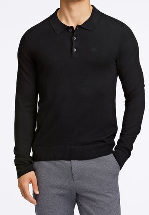 Mand iført en sort langærmet polo med tre knapper og grå bukser, stående med den ene hånd i lommen.