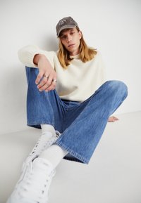 Romige chunky trui, blauwe denim jeans, witte sneakers en een grijze pet. Model zittend met gekruiste benen, waarbij ringen en textuur op de stoffen worden getoond.