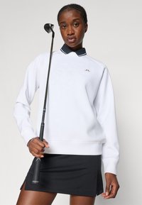 Biely sveter s čiernym golierom a zlatým logom, skombinovaný s čiernou sukňou. Model drží golfové palo s textúrovaným uchopením.