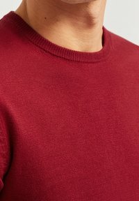 Czerwony sweter pleciony z okrągłym dekoltem. Tkanina jest gładka o delikatnej strukturze, z ribbowanym wykończeniem u szyi i mankietów.