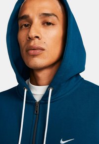 Tealfärgad zip-up hoodie med dragskohuva, vita detaljer och en liten Nike-logotyp på bröstet, tillverkad av mjukt material med en slät textur.