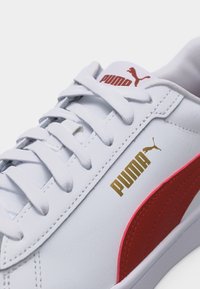 Λευκό αθλητικό παπούτσι Puma με κόκκινη πλευρική ρίγα, χρυσό λογότυπο Puma στο πλάι και λευκά κορδόνια δεμένα τακτικά.