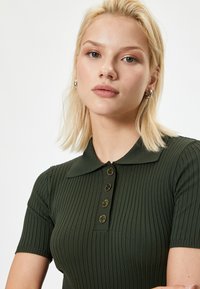 Polo-shirt vert à rayures, avec des manches courtes, une patte de boutonnage et un col plat. Texture lisse et design ajusté.